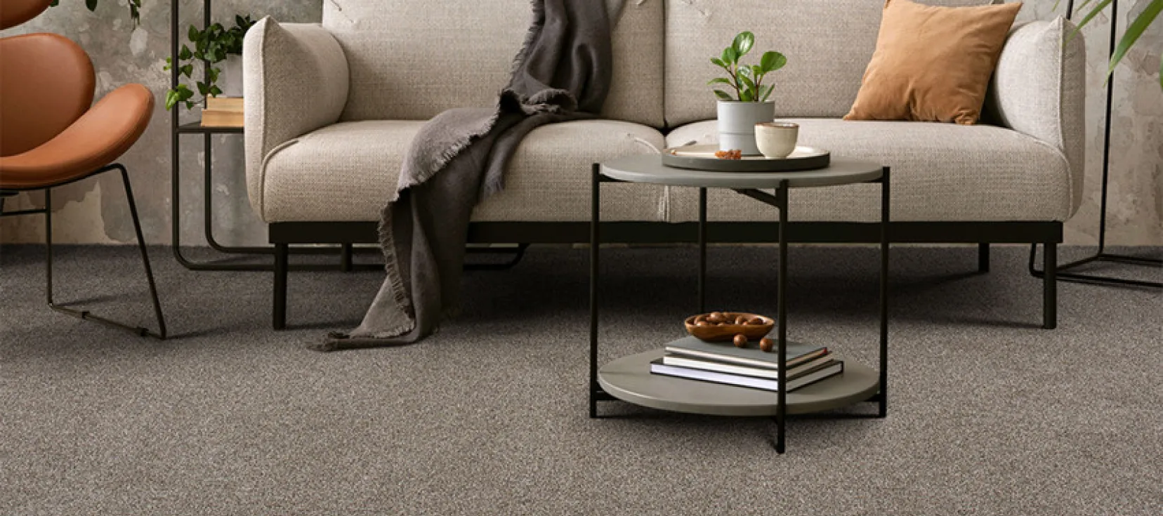 A stylish living room featuring Engineered Floors Carpet in a dark gray tone. A beige sofa with pillows and a gray throw is paired with a modern circular coffee table, books, and decor items.